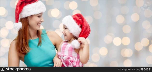christmas, family, childhood and people concept - happy mother and little girl in santa hats over holidays lights background