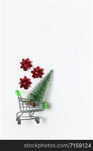 Christmas fir and red bows for gift in toy shopping cart on white background, copy space. New Year, sales, online shopping concept. Vertical, flat lay. Minimal style. Top view. For social media.