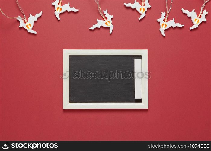 Christmas garland in form of white running deers, small chalk board with copy space on dark red background. Blank for postcard layout. Festive, New Year concept. Horizontal, flat lay. Minimal style.