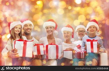 christmas, generation, holidays and people concept - happy family in santa helper hats with gift boxes sitting on couch over lights background