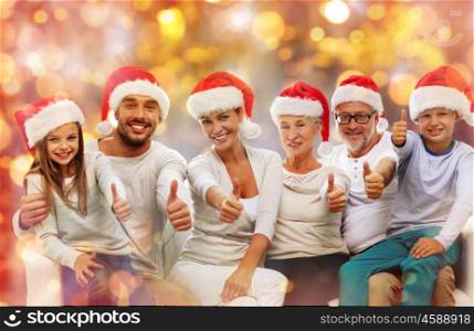 christmas, generation, holidays and people concept - happy family in santa helper hats sitting on couch and showing thumbs up gesture over lights background