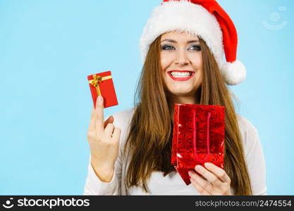 Christmas gifts. Happy woman wearing santa claus hat holding present red bag and small gift box with jewelery, on blue. Happy Christmas woman holds red gift box