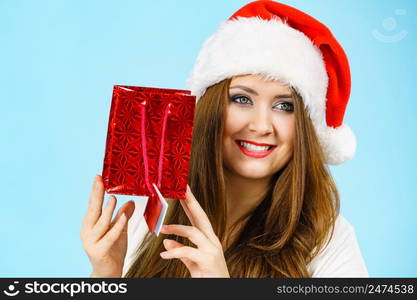 Christmas gifts. Positive young female wearing santa claus hat holding present red gift bag, on blue. Happy Christmas woman holds red gift bag