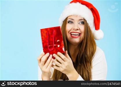 Christmas gifts. Positive young female wearing santa claus hat holding present red gift bag, on blue. Happy Christmas woman holds red gift bag