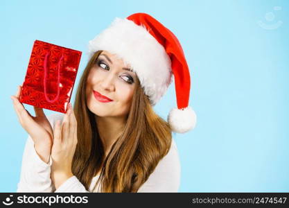 Christmas gifts. Positive young female wearing santa claus hat holding present red gift bag, on blue. Happy Christmas woman holds red gift bag