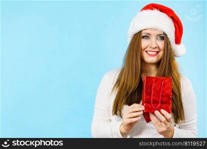 Christmas gifts. Positive young female wearing santa claus hat holding present red gift bag, on blue. Happy Christmas woman holds red gift bag
