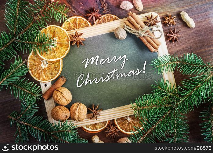 Christmas greeting card: chalkboard with inscription Merry Christmas! surrounded by green fir branches, nuts, oranges and spices