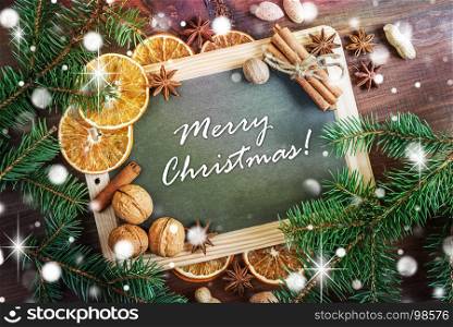 Christmas greeting card: chalkboard with inscription Merry Christmas! surrounded by green fir branches, nuts, oranges and spices