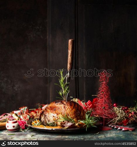 Christmas ham with stuck fork and rosemary on table with festive holiday decoration at wooden background, side view, place for text