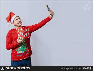 Christmas handsome man taking a selfie holding a gift isolated. Happy guy taking a christmas selfie isolated, Smiling young man in christmas hat holding a gift and taking a selfie isolated
