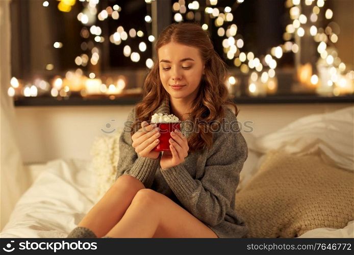 christmas, holiday and people concept - happy young woman in pullover holding mug with whipped cream in bed at night over festive lights on background. happy woman holding mug with whipped cream in bed