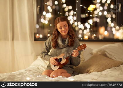 christmas, holiday and people concept - happy young woman playing ukulele guitar in bed at home bedroom at night over festive lights on background. happy young woman playing guitar in bed at home