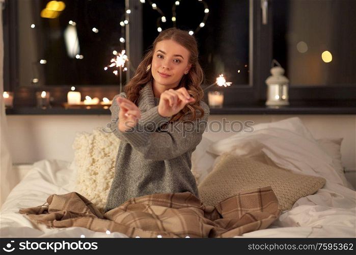 christmas, holiday and people concept - happy young woman with sparklers in bed at home bedroom at night. happy young woman with sparklers in bed at home