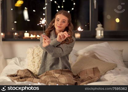 christmas, holiday and people concept - happy young woman with sparklers in bed at home bedroom at night. happy young woman with sparklers in bed at home