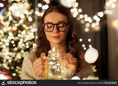 christmas, holiday and people concept - young woman in glasses with festive garland lights in mason jar mug at home. woman with christmas garland lights in glass mug