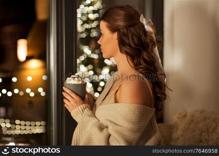christmas, holiday and people concept - young woman in pullover holding mug with whipped cream at window at night over festive lights on background. woman holding mug with whipped cream on christmas