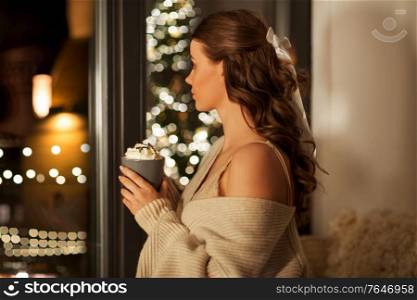 christmas, holiday and people concept - young woman in pullover holding mug with whipped cream at window at night over festive lights on background. woman holding mug with whipped cream on christmas