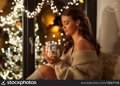 christmas, holiday and people concept - young woman in pullover sitting at window with festive garland lights in glass mason jar mug at home. woman with christmas garland lights in glass mug