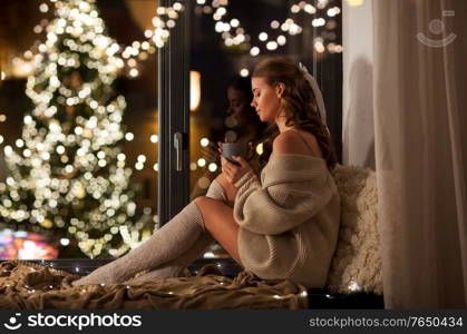 christmas, holiday and people concept - young woman in pullover sitting on windowsill with coffee or tea cup at home over festive lights on background. woman with cup of coffee on christmas at home