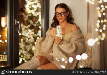christmas, holiday and people concept - young woman in sweater sitting on windowsill with festive garland lights in glass mason jar mug at home. woman with christmas garland lights in glass mug