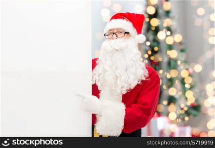 christmas, holidays, advertisement, gesture and people concept - man in costume of santa claus pointing finger to white blank billboard over tree lights background