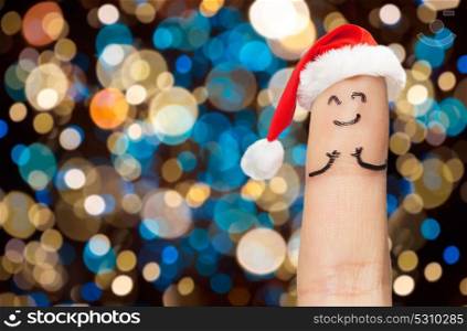 christmas, holidays and body parts concept - close up of one finger with smiley in santa hat over lights background. finger with smiley in santa hat at christmas
