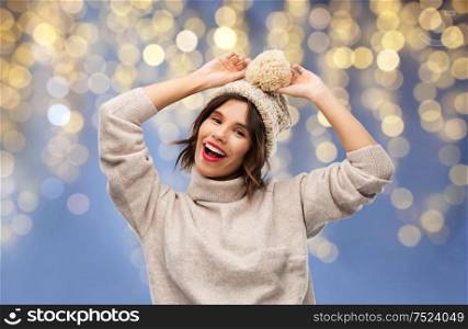 christmas, holidays and celebration concept - happy smiling young woman in knitted winter hat and sweater over festive lights on blue background. woman in winter hat and sweater on christmas