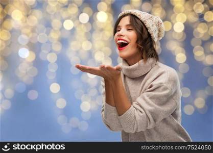 christmas, holidays and celebration concept - happy smiling young woman in knitted winter hat and sweater sending air kiss over festive lights on blue background. woman in winter hat sending air kiss on christmas
