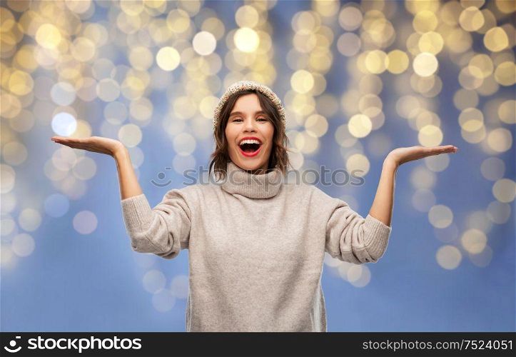 christmas, holidays and celebration concept - happy smiling young woman in knitted winter hat and sweater holding something on empty hand palm over festive lights on blue background. woman in winter hat holding something on christmas