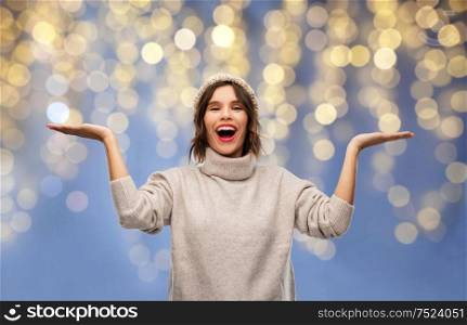 christmas, holidays and celebration concept - happy smiling young woman in knitted winter hat and sweater holding something on empty hand palm over festive lights on blue background. woman in winter hat holding something on christmas