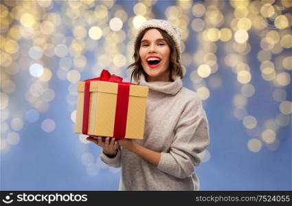 christmas, holidays and celebration concept - happy smiling young woman in knitted winter hat and sweater holding gift box over festive lights on blue background. young woman in winter hat holding christmas gift