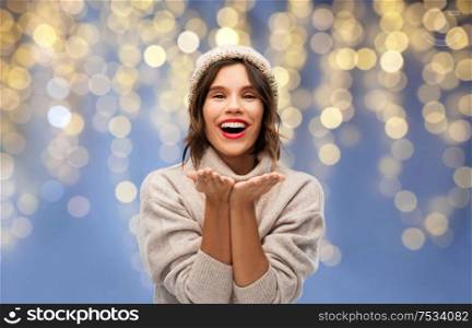 christmas, holidays and celebration concept - happy smiling young woman in knitted winter hat and sweater sending air kiss over festive lights on blue background. woman in winter hat sending air kiss on christmas