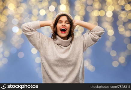 christmas, holidays and celebration concept - happy smiling young woman in knitted winter hat and sweater over festive lights on blue background. young woman in winter hat and sweater on christmas