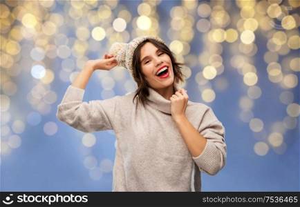 christmas, holidays and celebration concept - happy smiling young woman in knitted winter hat and sweater over festive lights on blue background. young woman in winter hat and sweater on christmas