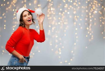 christmas, holidays and celebration concept - happy smiling young woman in santa helper hat over festive lights on grey background. happy young woman in santa hat on christmas