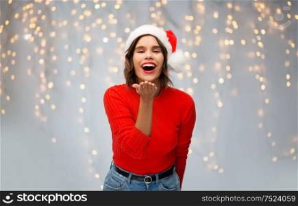christmas, holidays and celebration concept - happy smiling young woman in santa helper hat sending air kiss over festive lights on grey background. woman in santa hat sending air kiss on christmas