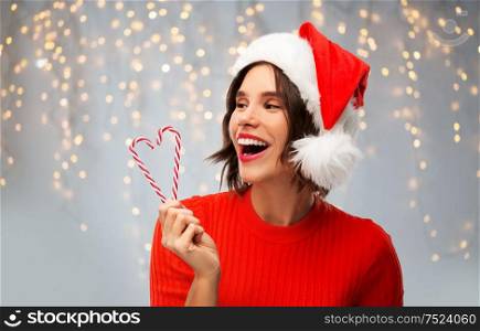 christmas, holidays and celebration concept - happy smiling young woman in santa helper hat with candy canes over festive lights on grey background. happy young woman in santa hat on christmas
