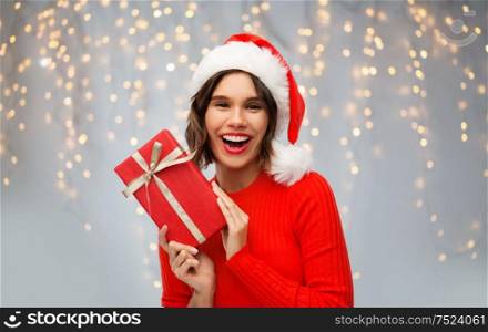christmas, holidays and celebration concept - happy smiling young woman in santa helper hat with red gift box over festive lights on grey background. happy woman in santa hat with red christmas gift