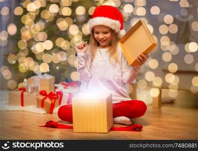 christmas, holidays and childhood concept - excited girl in santa helper hat opening gift box at home. excited girl in santa hat opening christmas gift