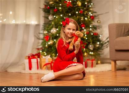 christmas, holidays and childhood concept - girl in red dress hugging teddy bear at home. girl in red dress hugging teddy bear at home