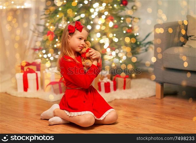 christmas, holidays and childhood concept - girl in red dress hugging teddy bear at home. girl in red dress hugging teddy bear at home