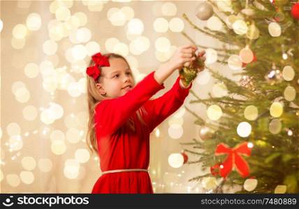 christmas, holidays and childhood concept - happy girl in red dress decorating natural fir tree. happy girl in red dress decorating christmas tree
