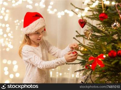 christmas, holidays and childhood concept - happy girl in santa hat decorating natural fir tree. happy girl in santa hat decorating christmas tree