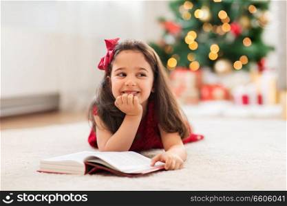 christmas, holidays and childhood concept - happy little girl lying on floor and reading book at home. happy girl reading book at home on christmas