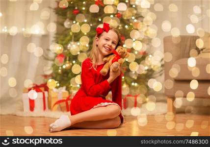 christmas, holidays and childhood concept - smiling girl in red dress hugging teddy bear at home. girl in red dress hugging teddy bear at home