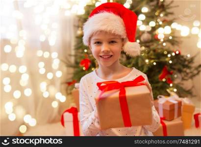 christmas, holidays and childhood concept - smiling girl in santa helper hat with gift box at home. smiling girl in santa hat with christmas gift