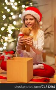christmas, holidays and childhood concept - smiling girl in santa helper hat with gift box and teddy bear at home. smiling girl in santa hat with christmas gift