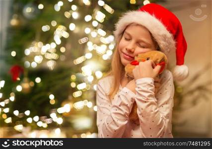 christmas, holidays and childhood concept - smiling girl in santa helper hat with gift box and teddy bear at home. smiling girl in santa hat with christmas gift