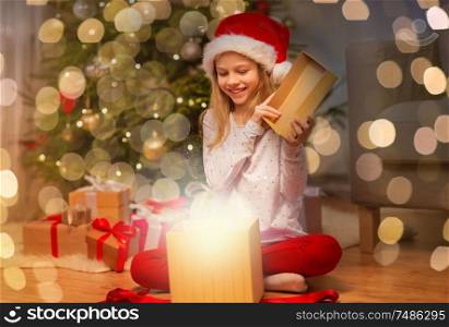 christmas, holidays and childhood concept - smiling girl in santa helper hat opening gift box at home. smiling girl in santa hat opening christmas gift