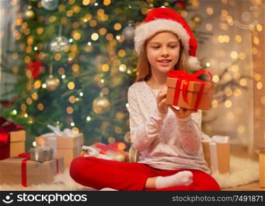 christmas, holidays and childhood concept - smiling girl in santa helper hat with gift box at home. smiling girl in santa hat with christmas gift